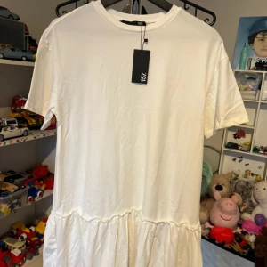 Vit t-shirtklänning från 157 - Enkel och snygg vit t-shirtklänning från 157 med rund halsringning och kort ärm. Klänningen har en avslappnad passform och volang nedtill som ger en cool och trendig look. Perfekt för dig som gillar en clean och stilren vibe.