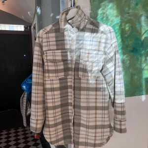 Rutig overshirt från KappAhl XS - Säljer en rutig overshirt från KappAhl i storlek XS. Jackan har grå, beige och vita toner med inslag av blått och brunt, två bröstfickor med lock och knappar framtill. Materialet är mjukt och känns som fleece, perfekt för lager på lager. Långärmad och med klassisk krage.