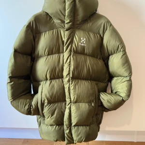 Grön pufferjacka från Haglöfs S - Säljer en grön pufferjacka från Haglöfs i storlek S. Jackan har huva, två stora fickor och quiltad design med breda paneler. Materialet är syntet och jackan har dragkedja framtill. Perfekt för kalla dagar och har en clean, sportig look.