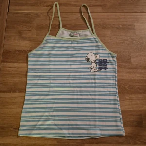 Randig Snoopy linnetopp i blått och vitt - Supersöt randig linnetopp från Snoopy med tunna axelband och tryck av Snoopy på bröstet. Toppen har blå och vita horisontella ränder och ljusgröna kantband. Perfekt för en chill sommarlook! Varan är helt oanvänd med etiketten kvar.
