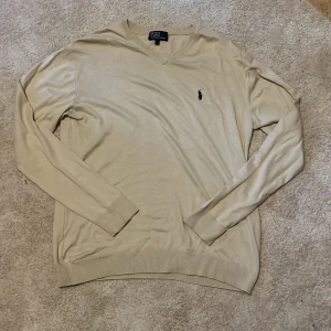 Beige tröja från Polo Ralph Lauren - Snygg beige tröja från Polo Ralph Lauren med v-ringning och broderad logga på bröstet. Långärmad modell i mjukt material, perfekt för lager-på-lager. Klassisk och stilren design som funkar till det mesta.