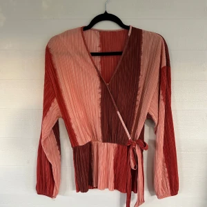 Randig omlottopp i rött och rosa - Säljer en unik omlottopp med plisserat tyg i röd- och rosatonade ränder. Toppen har långa ärmar, v-ringning och knytband i midjan. Perfekt för dig som gillar färg och coola detaljer!🤩💕