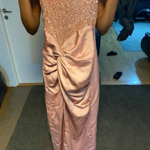 Glittrig rosa långklänning med slits - Säljer en superfin långklänning i ljusrosa med glittrig överdel och axelbandslös design. Kjolen är i satinliknande material med snygg drapering och stor rosett framtill samt hög slits. Perfekt för bal eller student! 💖priset kan diskuteras