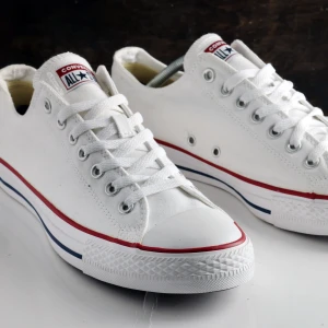 Vita Converse All Star sneakers - Klassiska vita Converse All Star sneakers i canvas med lågt skaft. Skorna har vita snören, metallfärgade öljetter och en vit sula med röda och blå ränder. Ikonisk logga på plösen och gummitå framtill. Perfekta för en clean och tidlös stil.