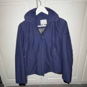 Blå vindjacka från J.Lindeberg - Snygg marinblå vindjacka från J.Lindeberg med dragkedja framtill och hög krage. Jackan har broderad logga på ärmen, justerbar nederkant och långa ärmar. Perfekt för blåsiga dagar och enkel att matcha med jeans. Perfekt nu till hösten🍁