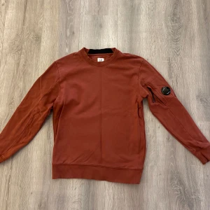  C.P. Company Sweatshirt  - Sweatshirt från C.P. Company med rund hals och ribbade muddar. Tröjan har en ikonisk linsdetalj på ärmen och en diskret ficka med dragkedja. Tillverkad i mjuk bomull, perfekt för en clean och stilren look.