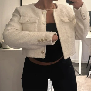 Vit croppad cardigan jacka med guldknappar - Säljer en vit croppad cardigan jacka med rund halsringning och dekorativa guldknappar framtill och på ärmsluten. Jackan har två bröstfickor med lock och är tillverkad i ett mjukt, strukturerat material som ger en lyxig känsla. Perfekt för att lyfta vilken outfit som helst.(pris går att diskuteras vid ett smidigt köp)
