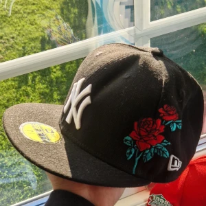 Svart New Era keps med rosor - Snygg svart keps från New Era med klassisk NY-logga framtill i vitt och broderade röda rosor med gröna blad på sidan. Flat skärm och gul storleksdekal kvar på skärmen. Perfekt för dig som vill sticka ut med en unik streetstyle.