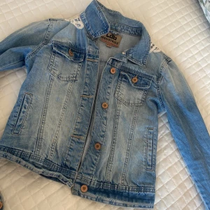 Ljusblå jeansjacka med spetsdetalj - Snygg ljusblå jeansjacka från Lab Industries med coola bronsfärgade knappar och två bröstfickor. Unik spetsdetalj i vitt på axlarna och ryggen som ger jackan en extra touch. Perfekt för dig som vill sticka ut med din stil.