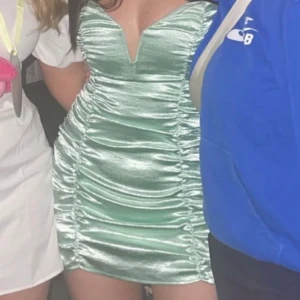 Mintgrön satin kortklänning - Snygg mintgrön kortklänning i satin med v-ringad hals och rynkade detaljer längs sidorna. Klänningen är figurnära och har en glansig finish som ger en lyxig känsla. 