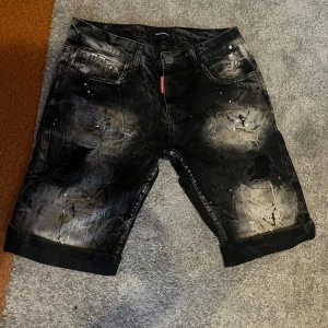 Svarta jeansshorts från Dsquared2 - Coola svarta jeansshorts från Dsquared2 med slitna detaljer, färgstänk och en gråvit tvättad effekt. Klassisk femficksmodell med uppvikta benslut och röd logotagg vid gylfen. Perfekta för dig som gillar streetstyle och edgy look.