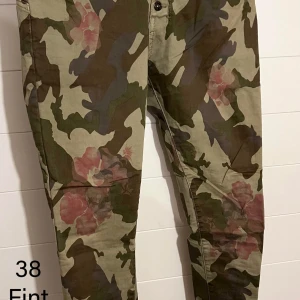 Byxor med camouflage och blommor - Säljer ett par byxor i camouflage-mönster med inslag av rosa blommor. Byxorna har klassisk passform och stängs med knapp och dragkedja. Perfekta för dig som vill sticka ut med en unik stil och gillar streetwear-vibbar.
