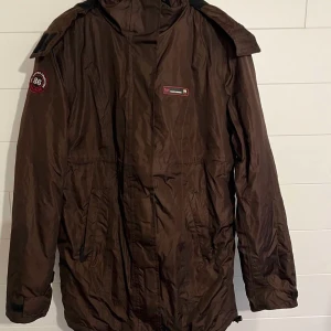 Brun parkajacka med huva - Säljer en brun vinter parkajacka från med huva och flera fickor. Jackan har patchdetaljer på ärmen och bröstet samt långa ärmar. Perfekt för kyligare dagar och har en klassisk parkamodell med dragkedja och knäppning framtill.