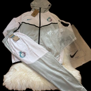Nike tech set/tracksuit al nassar limited edition - Nike hoodie och byxor i vitt och ljusgrått med svart dragkedja och huva. Tröjan har klubbmärke på bröstet och blå Nike-logga. Perfekt för träning eller chill, med sportig look och bekväm passform.