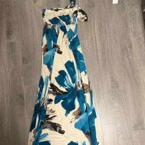 Blommig långklänning från 2Xtreme - Långklänning med stora blå och bruna blommor på beige botten från 2Xtreme. Klänningen har en oneshoulder-design med rynkade detaljer upptill och ett flowy fall. Perfekt för dig som vill sticka ut med en färgstark och unik look.
