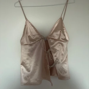 Champagnefärgad satinlinnetopp med knyt - Snygg champagnefärgad linnetopp i satin med smala axelband och öppen front som knyts med två band. Toppen har en glansig finish och en lätt croppad passform, perfekt för att skapa en trendig look.