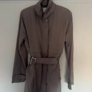 Beige trenchcoat med bälte från Lc wakiki  - Snygg beige trenchcoat med klassisk krage, bälte i midjan och slits bak. Jackan har långa ärmar med knappdetalj vid mudden och praktiska fickor. Perfekt för dig som gillar stilrena och tidlösa plagg.