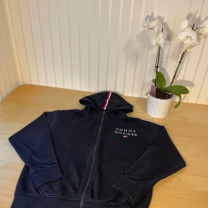 Marinblå hoodie från Tommy Hilfiger - Snygg marinblå hoodie från Tommy Hilfiger med dragkedja och huva. Klassisk logga på bröstet och en röd, vit och marinblå rand längs huvan. Mjuk bomullskänsla och ribbade muddar vid ärmslut och midja. Perfekt för chill dagar.