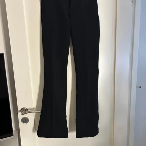 Kostymbyxor Vilma - Snygga svarta byxor med utsvängda ben och medelhög midja. Byxorna har pressveck, sidfickor och dekorativa bakfickor. Tillverkade i stretchigt material av återvunnen polyester och elastan för en bekväm passform. Perfekta för en trendig och stilren look. Knappt använda. Köpta för 499kr