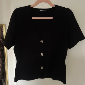 Topp från Gina Tricot - Svart topp från Gina Tricot med korta ärmar och v-ringning. Toppen har unika silvriga knappar framtill och en extra knapp medföljer. Modellen är figursydd och passar perfekt till jeans eller kjol för en clean look.