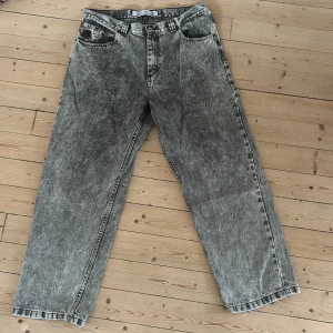 Acid wash 93! Jeans  - Säljer ett par gråsvarta acid wash jeans från 93! med bred passform och råa, fransiga benslut. Jeansen har klassisk femficksdesign, hög midja och en gul patch med graffiti-tryck bak. Perfekta för dig som gillar streetstyle och vintagevibbar.