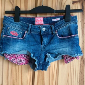 Superdry jeansshorts - Snygga blå jeansshorts från Superdry med råa kanter och rosa paisleymönstrat innerfoder. Shortsen har låg midja, klassiska fickor och coola rosa detaljer vid fickorna. Perfekta för sommaren och ger en avslappnad vibe.