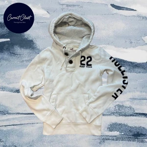 Vit hoodie från Hollister med tryck - Vit hoodie från Hollister med svart tryck på ärmen och bröstet. Tröjan har huva med snörning, knappar vid halsen och en stor magficka | Size S | Längd: 61cm Axelbredd: 42cm