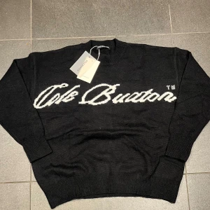 Svart stickad tröja Cole Buxton - Svart stickad tröja från Cole Buxton med vit logotyp i stor stil över bröstet. Klassisk rund halsringning och ribbade muddar vid ärmslut och nederkant. Perfekt för dig som gillar streetwear och vill ha en clean look.