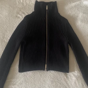 Svart ribbad kofta med dragkedja - Säljer en svart ribbad kofta med hög krage och hel dragkedja framtill. Tröjan är stickad och har långa ärmar, perfekt för kyligare dagar. Enkel och stilren design som passar till det mesta.
