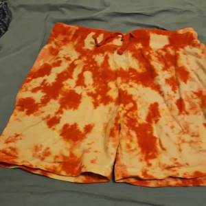Orange tie-dye shorts från TIKI - Snygga shorts från TIKI i orange och vitt tie-dye-mönster. De har en bekväm resårmidja med snörning och är tillverkade i mjuk manchester. Perfekta för chill dagar och sommarhäng.