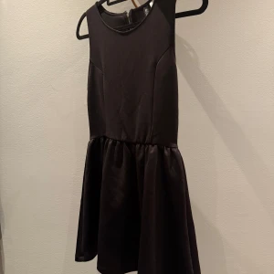 Svart ärmlös kort klänning H&M - Supersnygg svart klänning från Divided/H&M med ärmlös design och rund halsringning. Kjolen är lätt utställd och har en markerad midja. Klänningen har en synlig dragkedja i ryggen och är tillverkad i ett slätt och glansigt material.