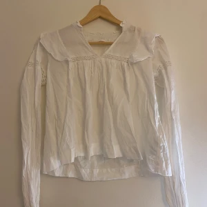 Vit blus med spets och volangdetaljer - Inte använd många gånger, fint skick. Stl 158/164. Säljer en romantisk vit blus i bomull med långa ärmar, spetsinslag och volangdetaljer vid axlarna. Blusen har v-ringning och en lös passform som ger en avslappnad men ändå söt look. Perfekt för dig som gillar vintage vibes och klassiska detaljer. 