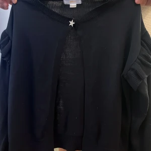 Svart kofta med stjärnknapp Gina Tricot - Svart kofta från Gina Tricot med långärmad design och volangdetaljer på ärmarna. Koftan har en snygg stjärnformad knapp upptill och ribbade muddar. Perfekt för dig som vill ha en enkel men ändå unik look. Har bara använt på ett skolfoto