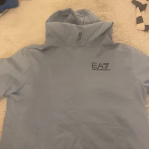 Ljusblå hoodie från EA7 Emporio Armani - Säljer en stilren ljusblå hoodie från EA7 Emporio Armani med logga på bröstet. Tröjan har huva och en stor magficka framtill.