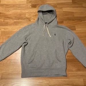 Grå hoodie från Polo Ralph Lauren - Klassisk grå hoodie från Polo Ralph Lauren med broderad marinblå logga på bröstet. Tröjan har huva med vita snören och en stor magficka. Mjuk och skön bomullsblandning, perfekt för chill dagar. Passformen är normal och ärmarna är långa. Det står M men sitter mer som S. Den har lite defekter som knoppning och en fläck 