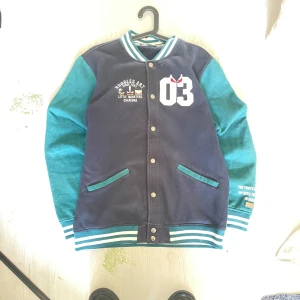 Blågrön bomberjacka Huggles Art - Snygg bomberjacka i blått och grönt från Huggles Art. Jackan har vita detaljer, broderad text och stort '03' framtill. Ribbade muddar med ränder, tryckknappar och två fickor. Materialet känns mjukt och är troligen polyester. Perfekt för dig som gillar streetstyle.