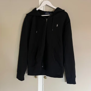 Svart hoodie från Polo Ralph Lauren - Svart hoodie från Polo Ralph Lauren med dragkedja framtill och klassisk logga broderad på bröstet. Tröjan har huva med snörning och två fickor framtill. Tillverkad i mjuk bomull och har en normal passform. Perfekt för en chill och stilren look.