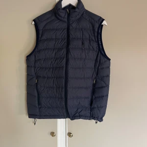 Mörkblå dunväst från Polo Ralph Lauren - Snygg mörkblå dunväst från Polo Ralph Lauren med broderad logga på bröstet. Västen har hög krage, dragkedja framtill och två fickor med dragkedja. Perfekt för lager-på-lager och har en clean, quiltad look.