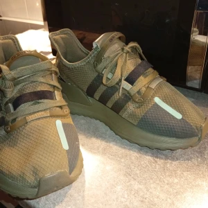  Sneakers i olivgrönt - Säljer ett par sneakers i olivgrönt med svarta detaljer och klassiska tre ränder på sidan. Skorna har mesh-överdel, snörning och en chunky sula för extra komfort. Perfekta för dig som gillar streetstyle och vill sticka ut med en unik färg.