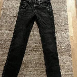 Svarta Replay Anbass jeans - Säljer ett par svarta Replay Anbass jeans med snygg tvätt och klassiska fem fickor. Jeansen har smal passform och raka ben, med diskreta slitningar och tydliga sömmar. Perfekta för dig som gillar en stilren och modern look.