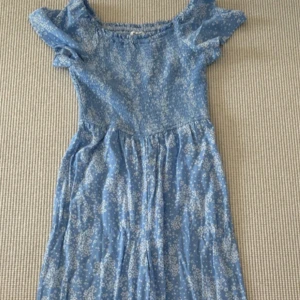 Blå blommig byxdress från Kappahl - Supersöt blå klänning från Kappahl med vita småblommiga mönster. Klänningen har korta puffärmar, smockad överdel och raka-vida jeans. Perfekt för varma dagar och har en romantisk vibe. 