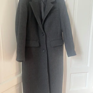 Grå ullkappa från H&M x Manteco, stl 34 - Snygg lång grå kappa från H&M i samarbete med Manteco, tillverkad i återvunnen ull. Klassisk rak modell med krage, två knappar framtill och fickor med lock. Perfekt för dig som gillar stilrena och tidlösa plagg.