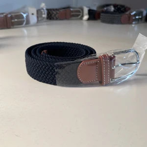 Mörkblått flätat bälte - Snyggt mörkblått bälte med flätad design och detaljer i brunt skinn vid spännet. Spännet är i silverfärgad metall och bältet har vita kontrastsömmar på skinnet. Perfekt accessoar för att lyfta din outfit. Bältet är elastiskt.