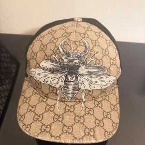 Gucci keps med insekt och GG-mönster - Säljer en beige och brun keps från Gucci med klassiskt GG-mönster och stor insekt-illustration framtill. Kepsen har svart mesh på baksidan och justerbart band med grönt och rött. Perfekt för dig som vill sticka ut med lyxig streetstyle.
