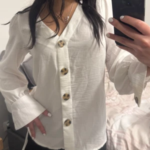 Vit blus med stora knappar - Säljer en vit blus med v-ringning och stora, mönstrade knappar framtill. Blusen har långa ärmar med breda manschetter och knappdetaljer. Den är tillverkad i ett lätt och luftigt tyg, perfekt för en stilren look.