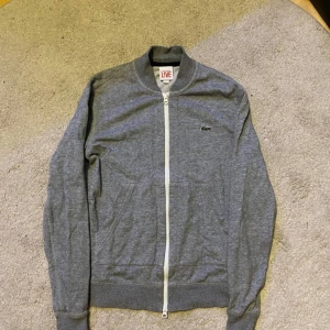 Lacoste grå ziptröja - Grå långärmad ziptröja från Lacoste LIVE med vit dragkedja och klassisk krokodillogga på bröstet. Tröjan har ribbade muddar och krage, perfekt för lager-på-lager. Tillverkad i mjuk bomull för en skön känsla.