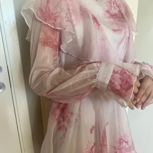 Ljusrosa blommig långklänning med volanger - Romantisk långklänning i ljusrosa med blommigt mönster i rosa toner. Klänningen har långa, transparenta ärmar med volangdetaljer och knyt i midjan. Volanger längs axlar och kjol ger en drömmig känsla. Perfekt för dig som gillar en feminin och bohemisk stil.