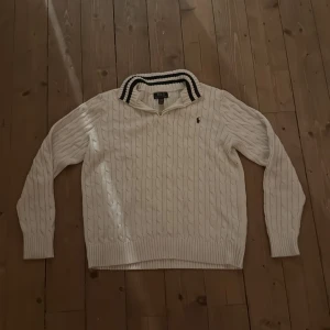 Vit kabelstickad tröja Polo Ralph Lauren - Kabelstickad vit tröja från Polo Ralph Lauren med klassisk krage och svarta ränder. Tröjan har lång ärm, ribbade muddar och dragkedja vid halsen. Ikonisk broderad logga på bröstet. Perfekt för en clean och sportig look.