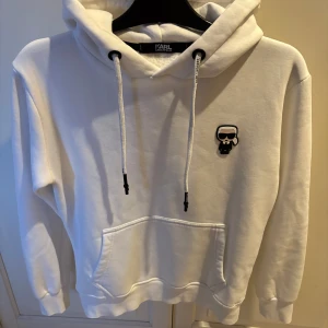 Vit hoodie från Karl Lagerfeld - Vit hoodie från Karl Lagerfeld med ikonisk Karl-brodyr på bröstet och logga på snörena. Mjuk insida, stor huva och känguruficka framtill. Snygg och clean design med svarta detaljer och tryck baktill vid nederkanten.