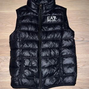 Svart dunväst från Emporio Armani EA7 - Stilren svart dunväst från Emporio Armani EA7 med quiltad design och hög krage. Västen har dragkedja framtill, två fickor och logga på bröstet. Perfekt för lager-på-lager perfekt till hösten. 